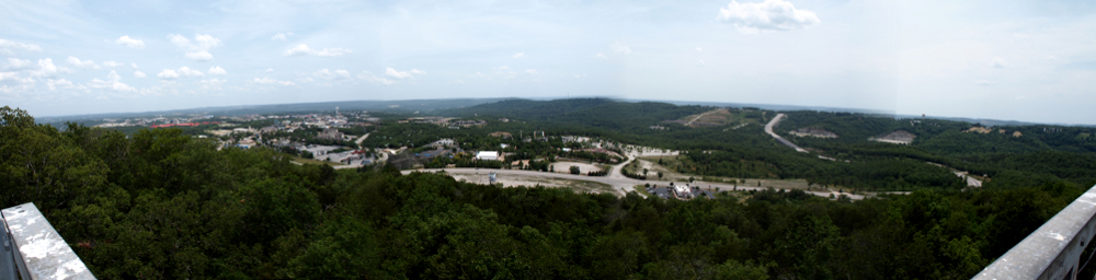 [Four photos stitched together giving a panoramic of green trees in foreground with buildings and road in middstanhe sky.]